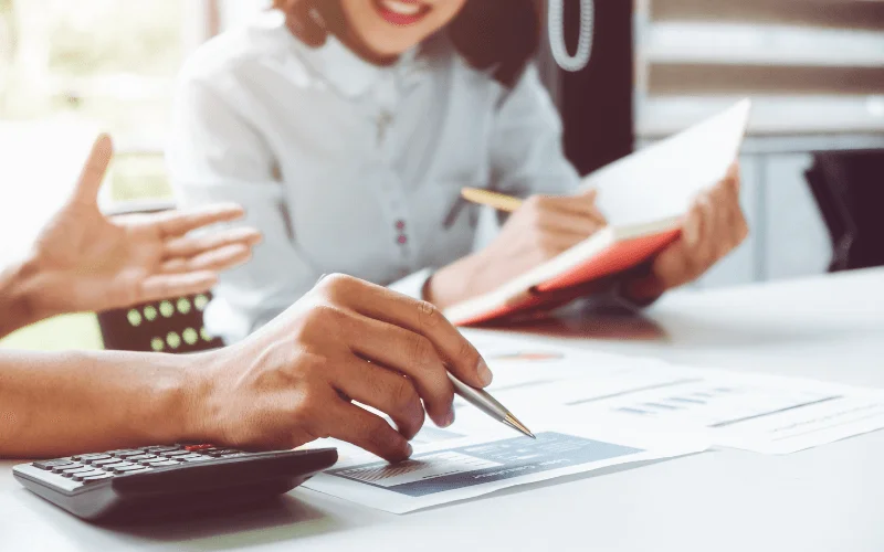 Manos revisando gráficos financieros y calculadora, mientras dos personas analizan estrategias para ampliación de capital online.